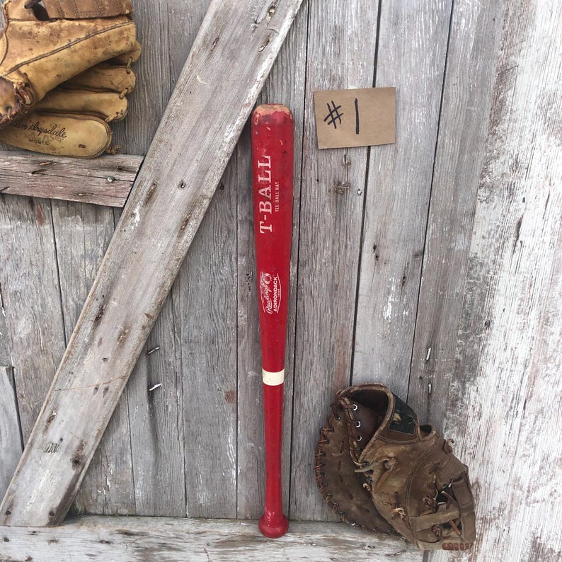 Puede incluir: Un bate de T-Ball rojo con detalles blancos y el texto "T-BALL THE BALL WAY" se muestra junto a un guante de b&eacute;isbol y una etiqueta que dice "#1". El bate es de Rawlings.