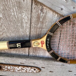May include: A vintage wooden tennis racket featuring a light tan frame with black accents and white stringing. The throat displays the Wilson logo, the text Champ, and a graphic of a trophy with three stars. The handle is wrapped in white tape.