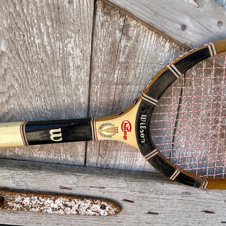 May include: A vintage wooden tennis racket featuring a light tan frame with black accents and white stringing. The throat displays the Wilson logo, the text Champ, and a graphic of a trophy with three stars. The handle is wrapped in white tape.