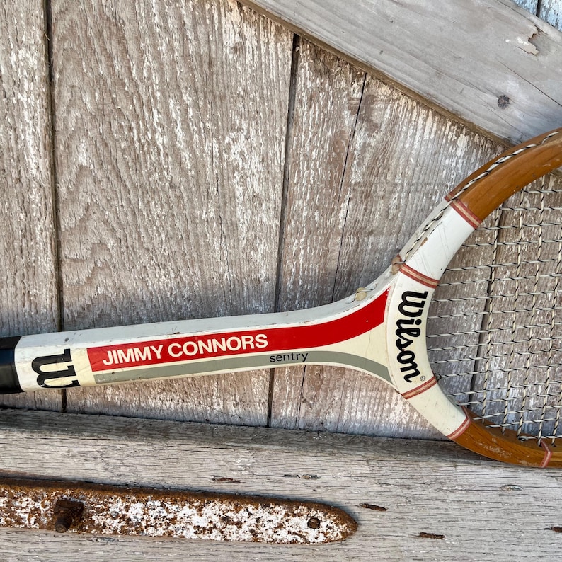 May include: A vintage Wilson Sentry tennis racket featuring a white frame with red and grey accents. The throat of the racket displays the text "JIMMY CONNORS" in red, "sentry" in black, and the Wilson logo in black on the white shaft.