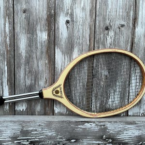 May include: A vintage wooden tennis racket with a cream-colored frame and a dark metal shaft. The racket head features a grid of nylon strings and a logo near the throat that reads TOURNEY with a laurel leaf design and a small circular emblem.