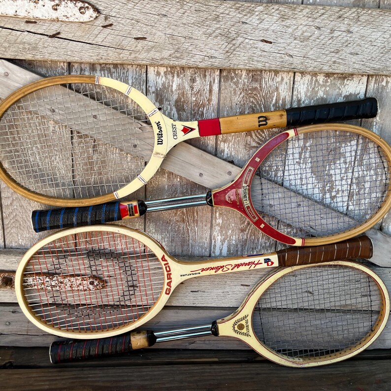 May include: Four vintage wooden tennis rackets with strung heads and wrapped handles. The rackets feature various brand names including Wilson Crest, Journey, Garcia Harold Solomon Signature, and another with a crest logo, all with metal shafts.
