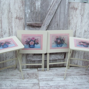 May include: Set of three white folding tables with floral designs. Each table features a different floral arrangement in a blue vase on a pink background.