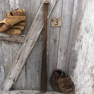 Puede incluir: Un juego vintage de bate, guante y guante de b&eacute;isbol sobre un fondo de madera desgastada. El bate es de color marr&oacute;n oscuro, y el guante y el guante son de cuero marr&oacute;n claro. Una peque&ntilde;a etiqueta con el n&uacute;mero "3" es visible.