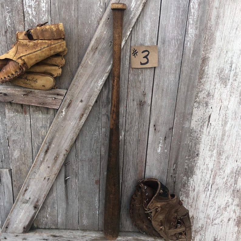 Puede incluir: Un juego vintage de bate, guante y guante de b&eacute;isbol sobre un fondo de madera desgastada. El bate es de color marr&oacute;n oscuro, y el guante y el guante son de cuero marr&oacute;n claro. Una peque&ntilde;a etiqueta con el n&uacute;mero "3" es visible.