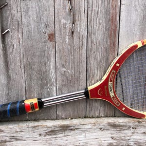 May include: A vintage tennis racket featuring a red and cream wooden frame with the text "TOURNEY-1" and "MacGregor" printed on the throat. The racket has a black grip with blue accents and a silver metal shaft connecting the handle to the head.