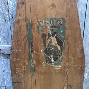 K&ouml;nnte beinhalten: Ein antikes, h&ouml;lzernes B&uuml;gelbrett mit einem verblassten Etikett, auf dem "To-Jid en-t Iron Folding Table" steht. Das Etikett zeigt eine Frau in einem Kleid beim B&uuml;geln.