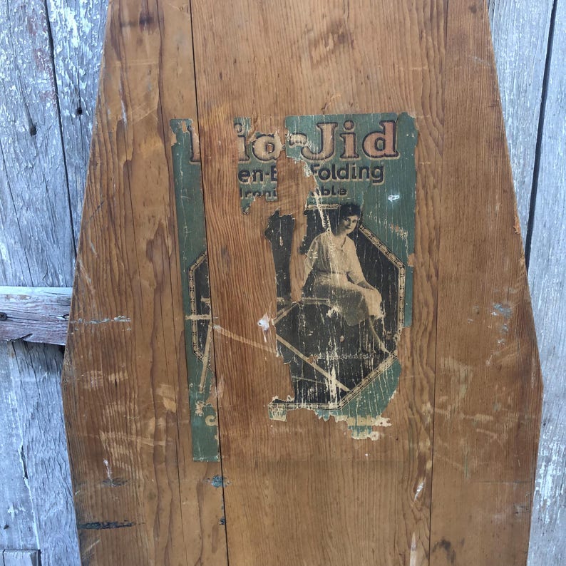 K&ouml;nnte beinhalten: Ein antikes, h&ouml;lzernes B&uuml;gelbrett mit einem verblassten Etikett, auf dem "To-Jid en-t Iron Folding Table" steht. Das Etikett zeigt eine Frau in einem Kleid beim B&uuml;geln.