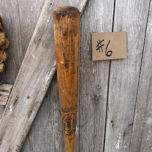 Puede incluir: Un bate de b&eacute;isbol Louisville Slugger vintage con el texto "George Brett" y "Louisville Slugger" impreso en la madera. El bate es de color marr&oacute;n claro con un aspecto desgastado. Tambi&eacute;n es visible una peque&ntilde;a etiqueta marr&oacute;n con el n&uacute;mero "6".