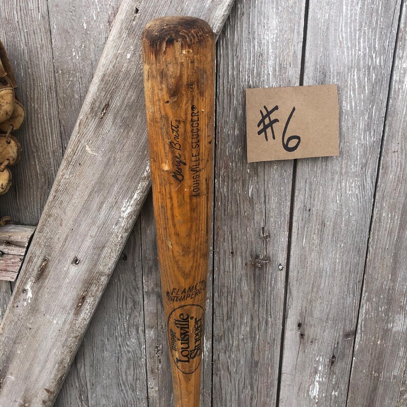 Puede incluir: Un bate de b&eacute;isbol Louisville Slugger vintage con el texto "George Brett" y "Louisville Slugger" impreso en la madera. El bate es de color marr&oacute;n claro con un aspecto desgastado. Tambi&eacute;n es visible una peque&ntilde;a etiqueta marr&oacute;n con el n&uacute;mero "6".