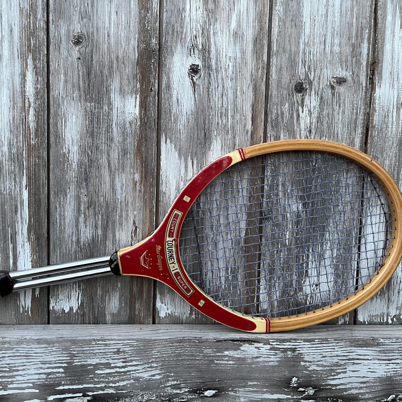 May include: A vintage wooden tennis racket featuring a natural wood frame with a red throat section. The red area displays gold text reading Tourney-1 Super and MacGregor with a crown logo. The racket has a metal shaft and dark nylon strings.
