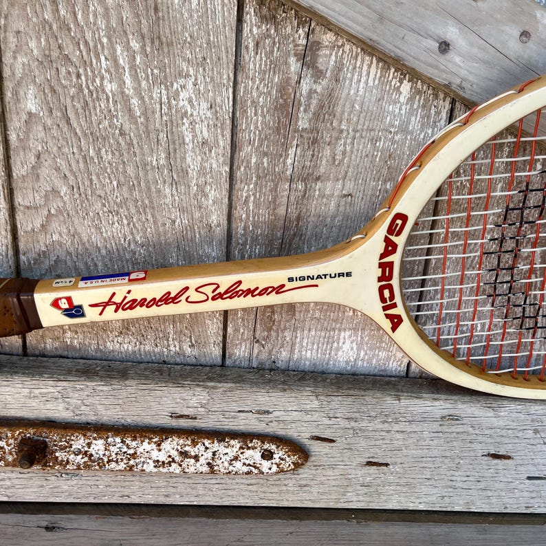 May include: A vintage wooden tennis racket featuring a cream-colored frame with red lettering that reads Garcia and Harold Solomon Signature. The racket includes white and red nylon strings and a small sticker indicating it was made in the USA.