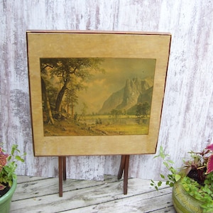 May include: A vintage wooden table with a landscape painting of a mountain scene. The table has three legs and is painted in a light brown color. The painting features a mountain range, trees, and a lake.