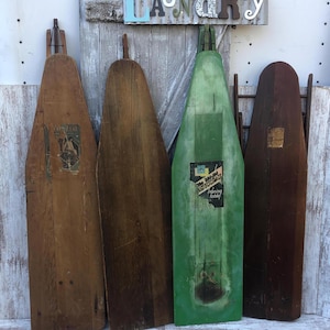 K&ouml;nnte beinhalten: Vier vintage B&uuml;gelbretter aus Holz, zwei sind braun und zwei sind gr&uuml;n. Die gr&uuml;nen Bretter haben Etiketten mit dem Text "The Perfect Ironing Board" und "Ironing Easily". Die Bretter lehnen an einer wei&szlig;en Wand mit einem Schild, auf dem "Laundry" in bunten Buchstaben steht.