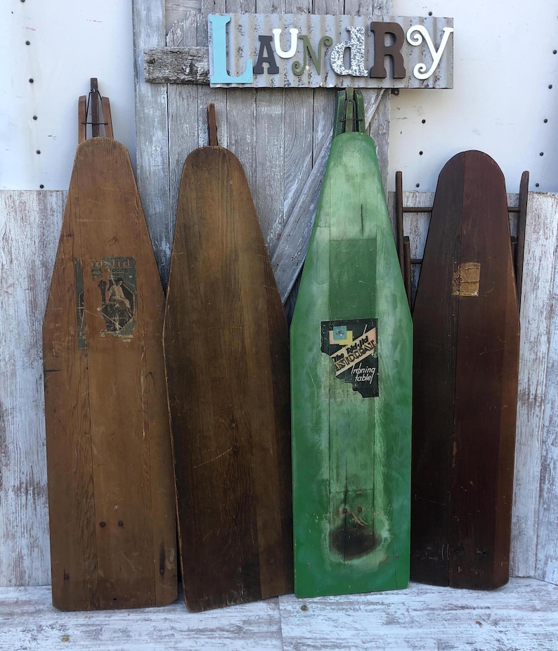 K&ouml;nnte beinhalten: Vier vintage B&uuml;gelbretter aus Holz, zwei sind braun und zwei sind gr&uuml;n. Die gr&uuml;nen Bretter haben Etiketten mit dem Text "The Perfect Ironing Board" und "Ironing Easily". Die Bretter lehnen an einer wei&szlig;en Wand mit einem Schild, auf dem "Laundry" in bunten Buchstaben steht.
