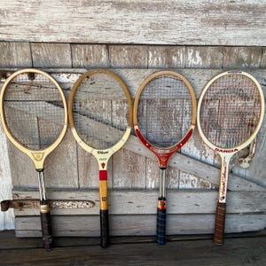 May include: Four vintage wooden tennis rackets arranged side by side. The first is light wood with a gold emblem, the second is a Wilson Crest with red accents, the third is a red and wood Tourney model, and the fourth is a Garcia with red strings.