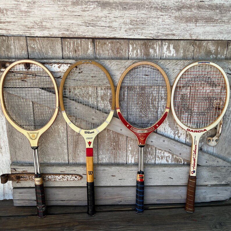 May include: Four vintage wooden tennis rackets arranged side by side. The first is light wood with a gold emblem, the second is a Wilson Crest with red accents, the third is a red and wood Tourney model, and the fourth is a Garcia with red strings.