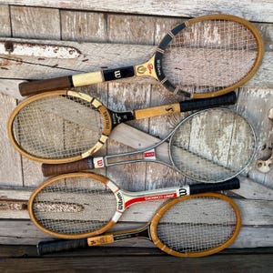 May include: A collection of five vintage tennis rackets featuring wooden and metal frames with nylon strings. The rackets include various brand names such as Wilson, Spalding, and Reindeer, with black, white, and brown grip tape on the handles.