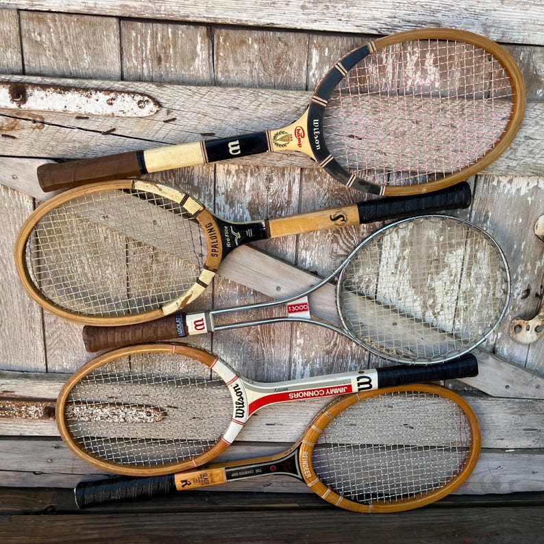 May include: A collection of five vintage tennis rackets featuring wooden and metal frames with nylon strings. The rackets include various brand names such as Wilson, Spalding, and Reindeer, with black, white, and brown grip tape on the handles.