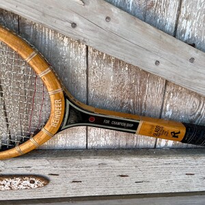 May include: A vintage wooden tennis racket with a light brown finish, featuring a black throat section with the text REINDEER and FOR CHAMPION SHIP. The frame includes a glass reinforced shaft label and a black grip at the handle end.
