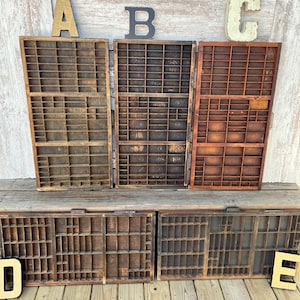 May include: Five rectangular wooden printer's trays with multiple small compartments arranged in a grid. Three trays are positioned vertically in the back row, and two trays are positioned horizontally in the front row, all featuring a dark brown finish.