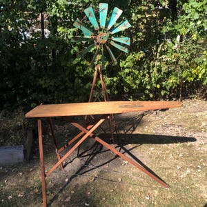 K&ouml;nnte beinhalten: Ein vintage B&uuml;gelbrett aus Holz mit einem klappbaren Metallgestell. Das Brett ist im Freien auf einem Rasen mit einem blauen und gr&uuml;nen Metallwindrad im Hintergrund aufgestellt.