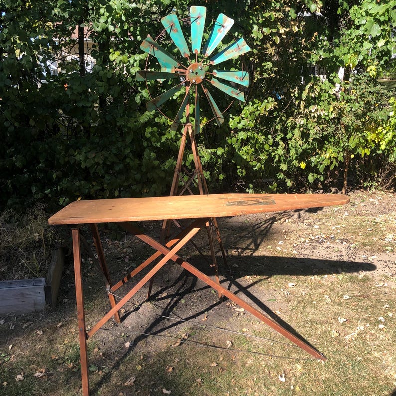 K&ouml;nnte beinhalten: Ein vintage B&uuml;gelbrett aus Holz mit einem klappbaren Metallgestell. Das Brett ist im Freien auf einem Rasen mit einem blauen und gr&uuml;nen Metallwindrad im Hintergrund aufgestellt.
