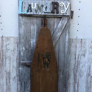 K&ouml;nnte beinhalten: Ein antikes, h&ouml;lzernes B&uuml;gelbrett mit einem Etikett, auf dem "To-Slid Easy Action" steht. Das Brett h&auml;ngt an einer verwitterten Holzt&uuml;r, &uuml;ber der das Wort "Laundry" in bunten Buchstaben geschrieben ist.