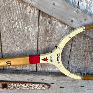 May include: A vintage Wilson Crest tennis racket featuring a natural wood handle with a black W logo, a red accent band, and a cream-colored throat section. The racket includes metal stringing and black text reading Wilson Crest with a red chevron.