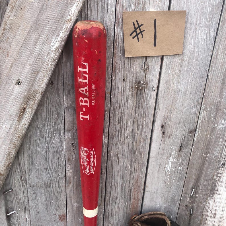 Puede incluir: Un bate de T-Ball rojo con letras blancas y una franja blanca cerca del mango. El bate tiene el logotipo de Rawlings Adirondack. Tambi&eacute;n es visible una peque&ntilde;a etiqueta marr&oacute;n con el n&uacute;mero "#1".