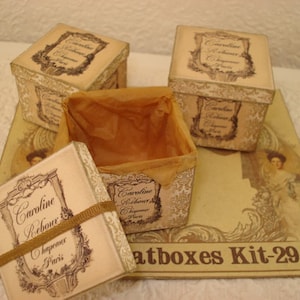 May include: A set of vintage-style decorative boxes with lids, featuring a cream-colored background and ornate labels. One box is open, revealing a brown interior. The boxes are arranged on a matching decorative base with the text "atboxes Kit-29".