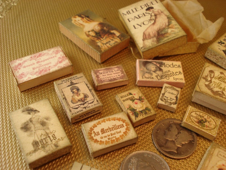 Peut inclure: Une collection de bo&icirc;tes miniatures de style vintage avec diff&eacute;rents motifs et textes. Les bo&icirc;tes sont en carton et ont un fond cr&egrave;me avec des lettres brunes et noires. Certaines bo&icirc;tes pr&eacute;sentent des images de femmes en v&ecirc;tements vintage, tandis que d'autres ont des textes tels que "Modes aux Elegantes" et "Mille Direts Paris Lyon".