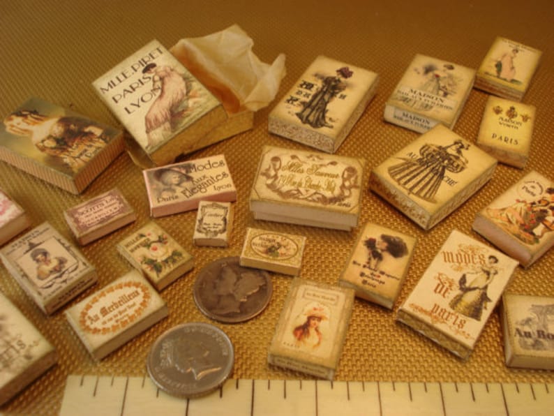 Peut inclure: Une collection de bo&icirc;tes miniatures avec des motifs et des textes vintage. Les bo&icirc;tes sont d&eacute;cor&eacute;es d'images de femmes en robes, de fleurs et d'autres &eacute;l&eacute;ments d&eacute;coratifs. Les bo&icirc;tes sont en carton et ont un aspect vieilli. Le texte sur les bo&icirc;tes comprend les mots "Paris", "Modes", "Elegantes" et "Au Merveilleux".