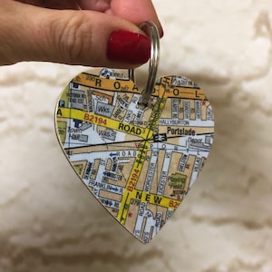 May include: A heart-shaped keyring with a map of a city printed on it. The map shows street names, roads, and landmarks. The keyring is attached to a silver ring.