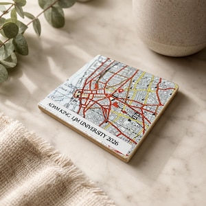 May include: A square ceramic coaster featuring a detailed city map with red and yellow street lines. A white rectangular label at the bottom displays the black text ADAM KING LJM UNIVERSITY 2026. The coaster has a smooth, glossy finish.