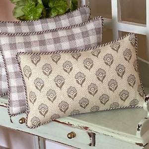 May include: Two rectangular throw pillows with a textured fabric finish. The front pillow features a repeating grey floral pattern on a beige background, while the back pillow has a grey and white gingham check pattern. Both pillows are edged with a twisted cord trim.