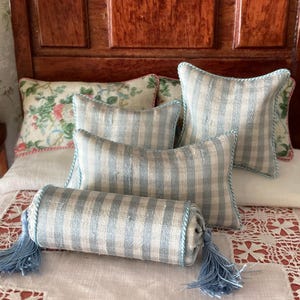 May include: Miniature bedding set featuring a bolster pillow with blue tassels and striped pillows in shades of blue and white. The set includes floral print pillows, all arranged on a white bed with a lace-trimmed cover.
