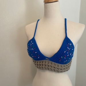 Puede incluir: Top de ganchillo azul con diseño halter. El top presenta un busto triangular azul con detalles bordados coloridos y una banda gris texturizada en la parte inferior. Se muestra en un maniquí. Medidas en centímetros.