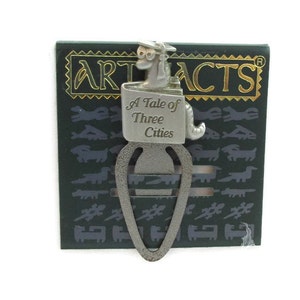 May include: A pewter bookmark featuring a cartoon dog wearing a graduation cap. The bookmark is shaped like a book and has the text "A Tale of Three Cities" engraved on it.