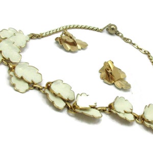 May include: A gold necklace and earrings set featuring white enamel leaves. The necklace has a chain with a gold ball clasp. The earrings are clip-on style.