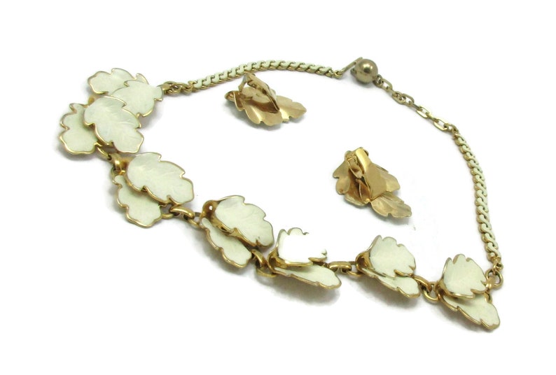 May include: A gold necklace and earrings set featuring white enamel leaves. The necklace has a chain with a gold ball clasp. The earrings are clip-on style.