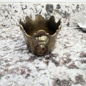 May include: A brass bowl with a scalloped, crown-like rim and a tapered body. The exterior features a decorative lion head motif on the front, which holds a circular metal ring. The metal surface has a weathered, aged patina with visible dark spots.
