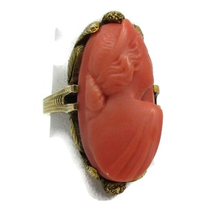 May include: A gold ring featuring a carved coral cameo of a woman's profile. The cameo is set in a gold frame with an intricate design.