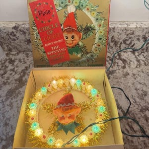 May include: A vintage Pixie Circle of Light spinning holiday light in its original box. The display features a central elf head surrounded by a circular gold tinsel wreath with alternating clear and teal miniature light bulbs and a green cord.