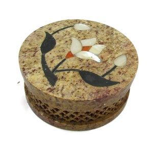 May include: A round, brown stone box with a carved floral design. The flower is made of white, orange, and black stone pieces. The box has a lattice pattern around the bottom.