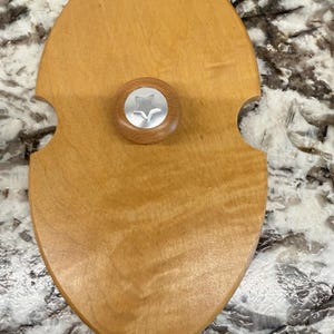 May include: An oval-shaped wooden press with a smooth, light brown finish and a visible wood grain pattern. The press features a central circular wooden knob topped with a silver-toned metal disc embossed with a five-pointed star design.