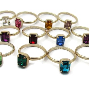 May include: Twelve gold rings with different colored gemstones. The gemstones are emerald green, light blue, dark blue, teal, light green, red, purple, pink, orange, and clear.