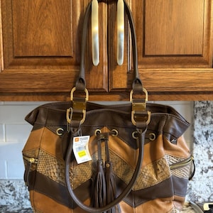 Pode incluir: Uma mala de mão em tons de castanho e bege com um padrão chevron que alterna painéis lisos e painéis com textura de pele de cobra. A mala inclui duas alças de ombro, ferragens douradas, bolsos laterais com fecho e uma borla decorativa.