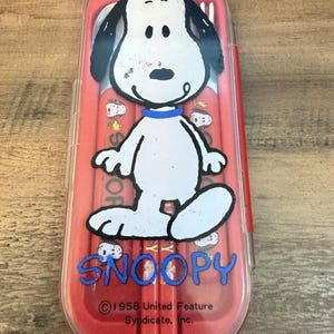 以下が含まれることがあります： 蓋に漫画キャラクターのスヌーピーの大きなグラフィックが描かれた、長方形の赤いプラスチック製カトラリーケース。ケースにはフォークと箸が入っており、表面には「SNOOPY」および「1958 United Feature Syndicate, Inc.」という文字が印刷されています。