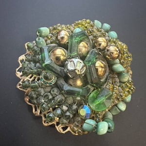 Puede incluir: Un broche vintage con un conjunto de cuentas verdes y doradas, cristales facetados y detalles metálicos. El broche presenta una variedad de formas y texturas, incluyendo cuentas redondas, cristales rectangulares y una base dorada de filigrana. El diseño general es ornamentado y llamativo.